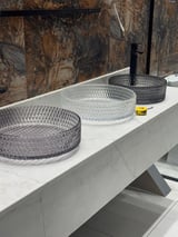 Three glass decorative trays displayed on a white marble bathroom vanity against a textured stone backsplash