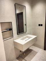 Modern bathroom vanity with white sink counter, black-framed mirror, and wall-mounted storage shelf on cream tile walls
